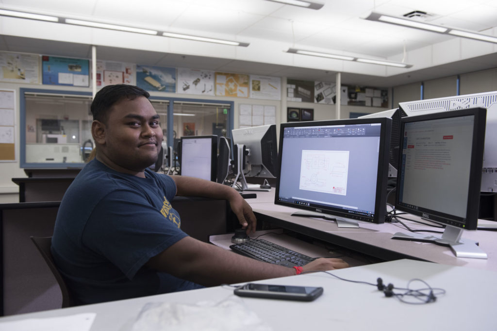 A male college student sits in front of a computer screen displaying CAD software.